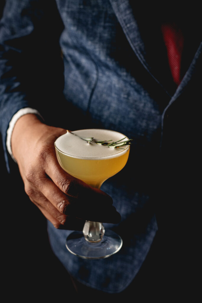 the King Charles cocktail with a sprig of rosemary on top and someone in a nice suit and red tie holding it.
