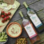 A bottle of Bee Lightly rose wine and a bottle of Bee Lightly sauvignon blanc, both made with 100% recycled plastic and lined with a proprietary gas barrier and UV inhibitor, on a table next to cheese and appetizers