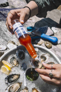 Hog Island Oyster Co Hot Sauce is shown driblling red dabs onto a round dish of oysters