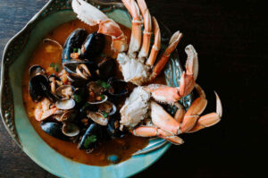 Taylor Shellfish Cioppino Kit cooks up to this – Crab and mussels emerging from a tomato -based sugo in a pale blue bowl
