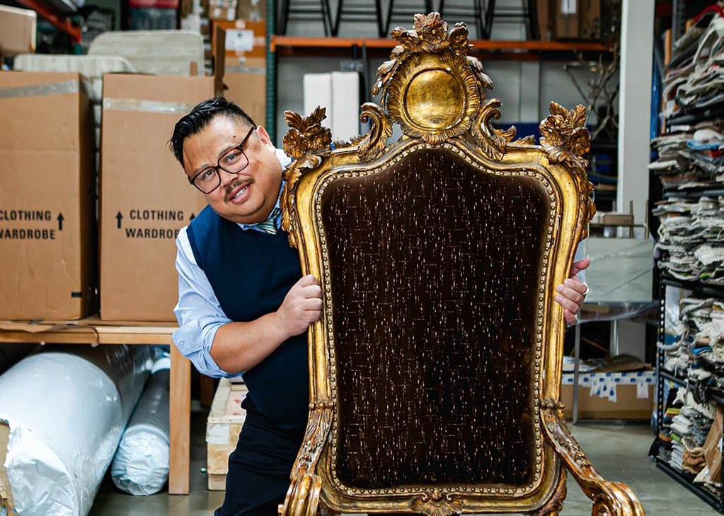 interior designer Jon de la Cruz looks at the camera from behind a sort-of kings' chair inside a warehouse