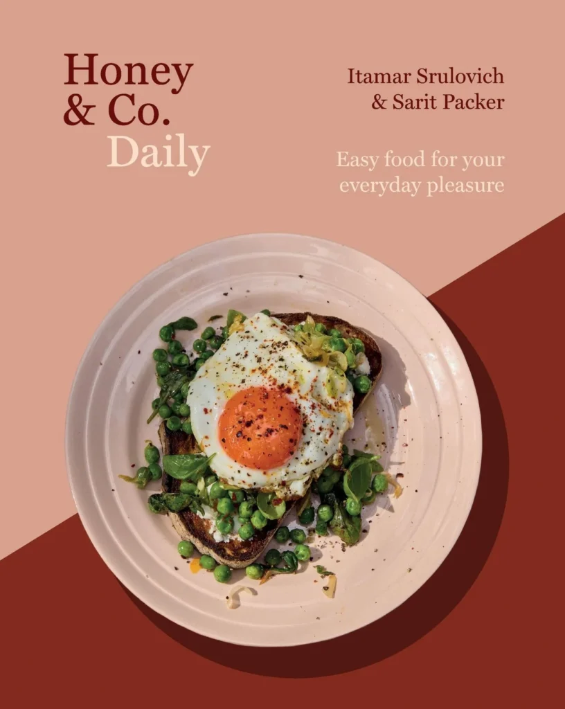 the cover of Honey & Co Daily cookbook is a white plate of toast and greens topped with an egg on top of a rust and dusty rose-colored cover