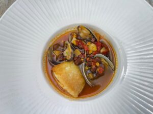 overhead view of halibut and clams in a light tomato and chorizo broth in a broad white bowl